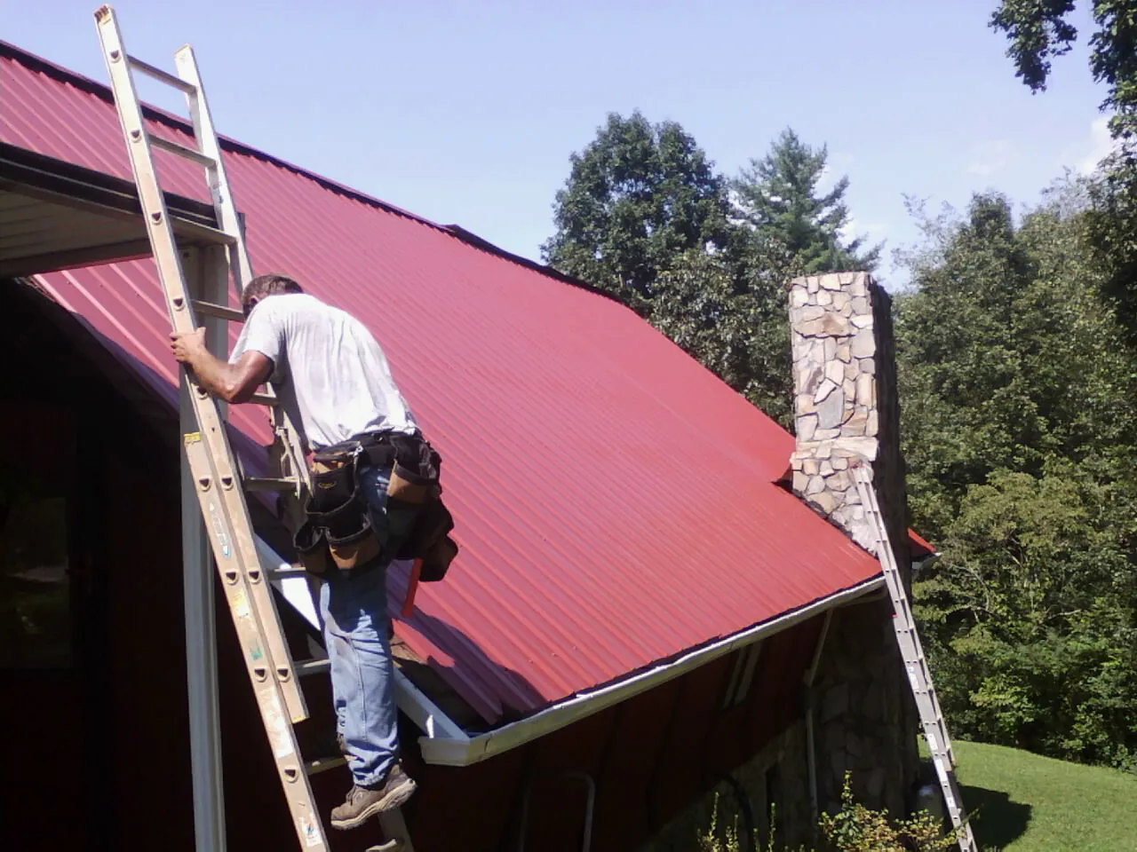 Roofer on a ladder beside a red metal roof and stone chimney — Soffit & Fascia Repair by Roofing Brothers in Penn Run